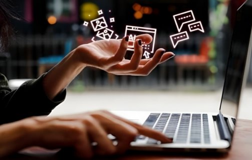 A imagem mostra uma mulher sentada em frente a um notebook e acima da suas mãos há hologramas de ícones da internet.