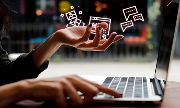 A imagem mostra uma mulher sentada em frente a um notebook e acima da suas mãos há hologramas de ícones da internet.