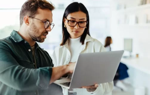A imagem mostra um homem e uma mulher conversando em um escritório. Ele está segurando um notebook e apontando algo para a tela, possivelmente dando a ela dicas de como saber se um site é seguro.