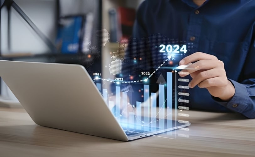 A imagem mostra um homem em frente a um notebook. Há um holograma acima do teclado com um gráfico em barras e mostra um aumento ao passar dos anos e o maior está no ano de 2024.