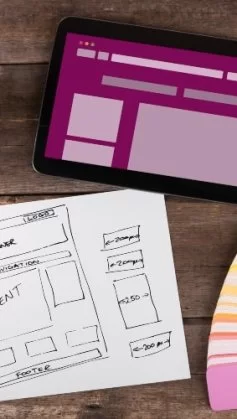 A imagem mostra rascunhos de telas de aplicativo desenhadas à mão, representando o processo de wireframe (etapa essencial do UX/UI design). Sobre a mesa estão canetas coloridas, notas adesivas e um notebook, simbolizando a fase criativa e analítica do desenvolvimento de produtos digitais. As setas entre as telas indicam o fluxo de navegação do usuário, demonstrando como ideias são estruturadas antes da implementação. Essa cena reflete o equilíbrio entre design funcional e experiência do usuário, fundamentais para criar interfaces intuitivas e eficientes.