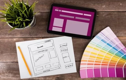 A imagem mostra rascunhos de telas de aplicativo desenhadas à mão, representando o processo de wireframe (etapa essencial do UX/UI design). Sobre a mesa estão canetas coloridas, notas adesivas e um notebook, simbolizando a fase criativa e analítica do desenvolvimento de produtos digitais. As setas entre as telas indicam o fluxo de navegação do usuário, demonstrando como ideias são estruturadas antes da implementação. Essa cena reflete o equilíbrio entre design funcional e experiência do usuário, fundamentais para criar interfaces intuitivas e eficientes.