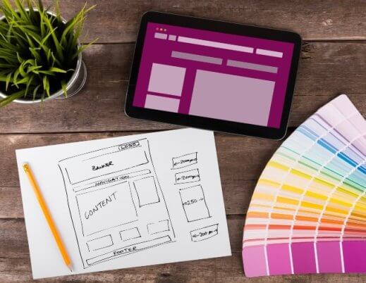 A imagem mostra rascunhos de telas de aplicativo desenhadas à mão, representando o processo de wireframe (etapa essencial do UX/UI design). Sobre a mesa estão canetas coloridas, notas adesivas e um notebook, simbolizando a fase criativa e analítica do desenvolvimento de produtos digitais. As setas entre as telas indicam o fluxo de navegação do usuário, demonstrando como ideias são estruturadas antes da implementação. Essa cena reflete o equilíbrio entre design funcional e experiência do usuário, fundamentais para criar interfaces intuitivas e eficientes.