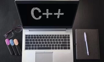 Laptop. Na tela, aparece os dizeres C++. Há canetas e um caderno ao redor, além de um par de óculos