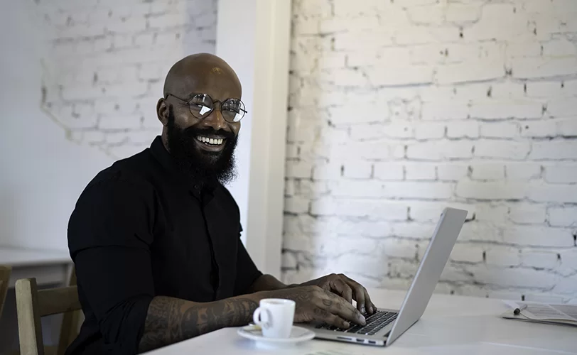Homem de óculos e barba em frente a um laptop. Há uma parede branca de tijolinhos atrás dele e uma xícara perto de sua mão