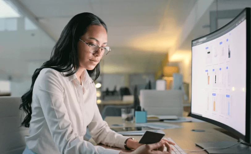 Mulher diante de um computador analisando algo que está nas mãos dela