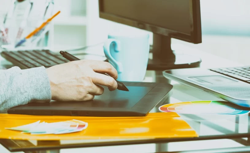 Pessoa manipulando um tablet com caneta diante de um desktop. Só aparece a mão dela