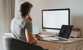 Homem trabalhando com um desktop, um laptop e um caderno diante dele. Ele está de costas para a câmera