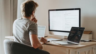 Homem trabalhando com um desktop, um laptop e um caderno diante dele. Ele está de costas para a câmera