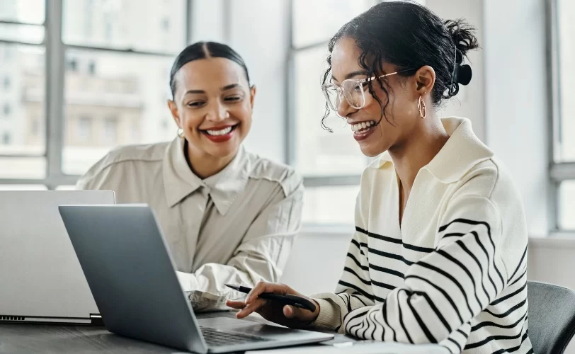 Duas mulheres trabalhando juntas em seus respectivos laptops. Elas sorriem olhando para a tela de uma delas