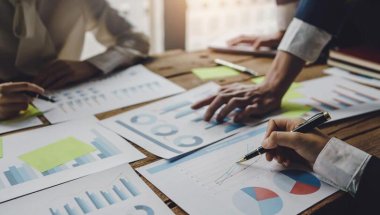 Pessoas analisando gráficos espalhados em papéis sobre uma mesa. Os rostos delas não aparecem