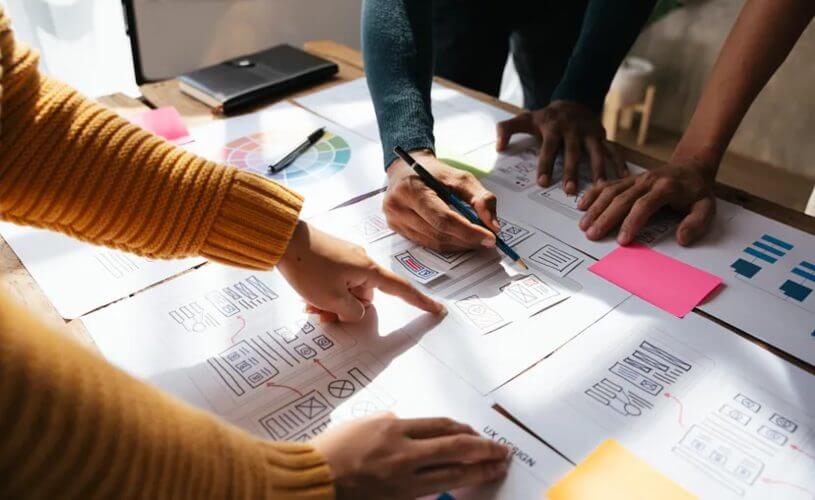 Pessoas apontando e desenhando em papéis sobre uma mesa que está entre elas