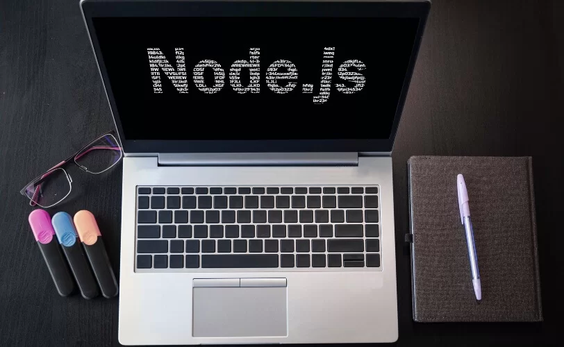 Notebook, canetas, caderno e óculos sobre superfície escura. Na tela do equipamento está escrito Node.Js