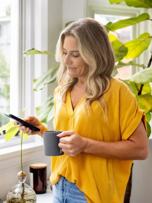 Mulher checando as redes sociais em seu celular e sorrindo. Ela segura uma caneca com a outra mão. Está perto de uma janela em um ambiente iluminado