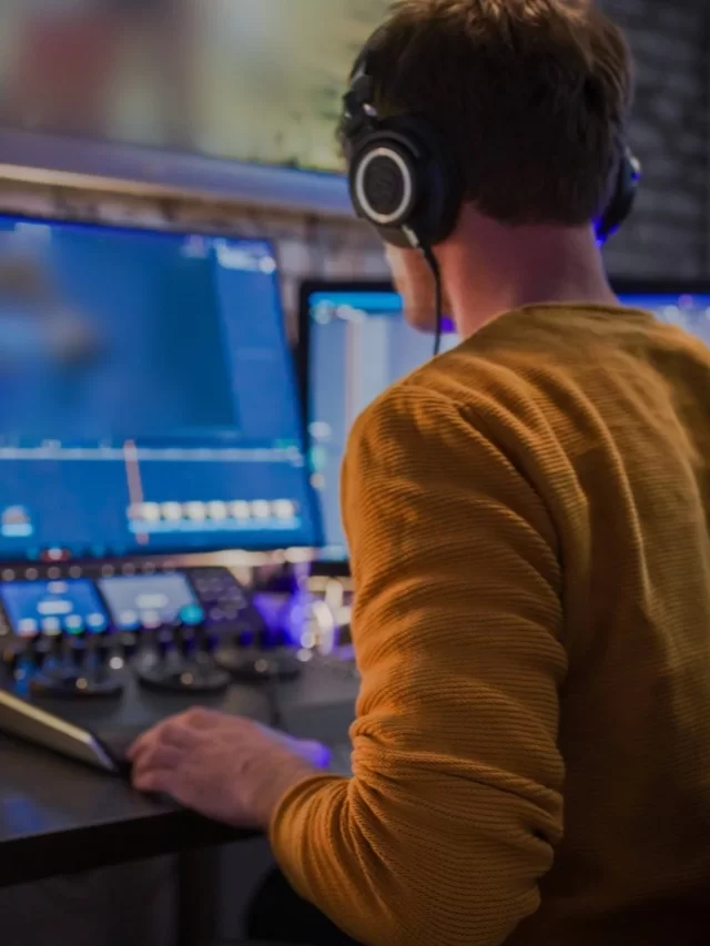 Homem jovem branco de cabelos curtos e escuros está de costas, editando músicas em seu estúdio usando a plataforma Suno AI. Ele está sentado em frente a três monitores, usando fones de ouvido e uma camiseta amarela de manga longa.