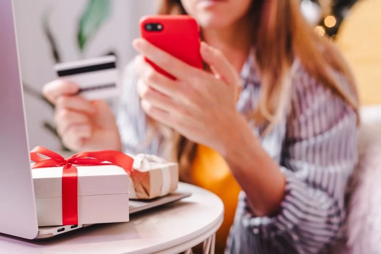 Mulher com cartão de crédito, realizando compra pelo smartphone e na mesa presentes em cima de um notebook.