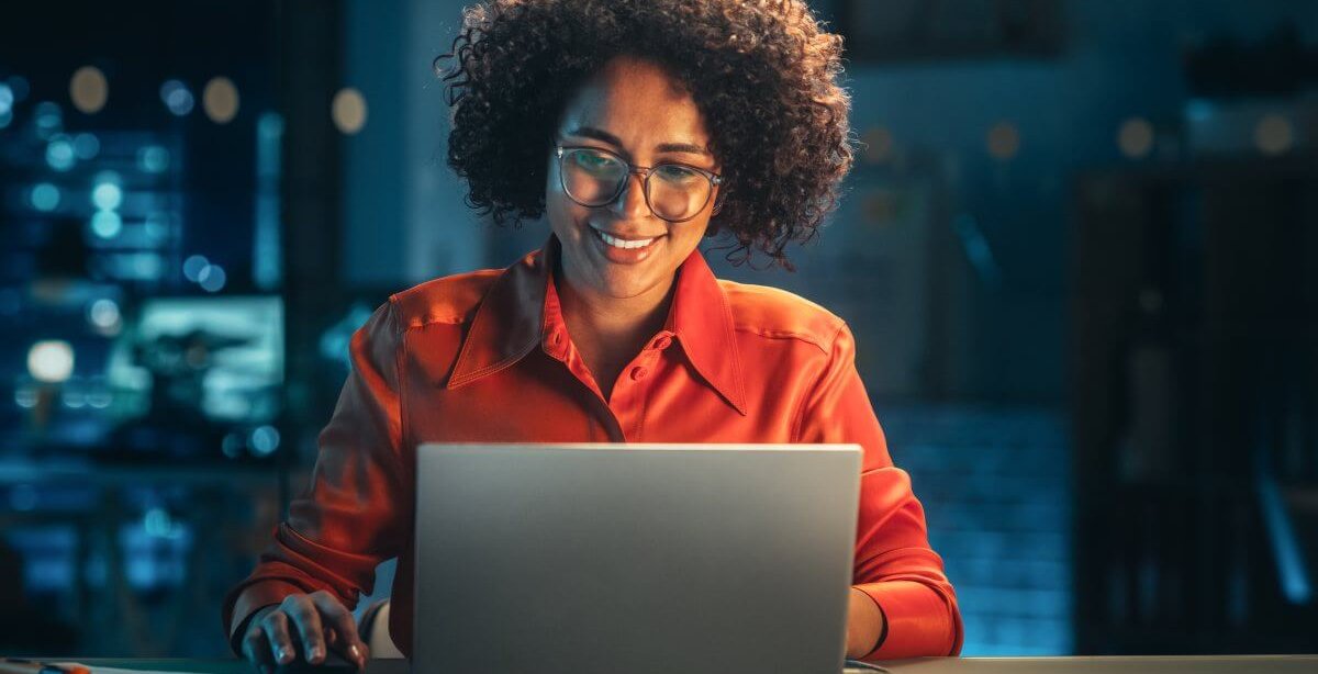 Mulher negra, usando óculos e camisa laranja, trabalha em um laptop e sorri. Atrás dela parece haver um servidor físico, mas ele está desfocado.