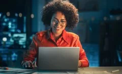 Mulher negra, usando óculos e camisa laranja, trabalha em um laptop e sorri. Atrás dela parece haver um servidor físico, mas ele está desfocado.