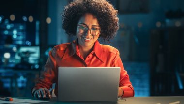 Mulher negra, usando óculos e camisa laranja, trabalha em um laptop e sorri. Atrás dela parece haver um servidor físico, mas ele está desfocado.