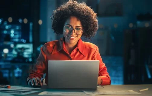 Mulher negra, usando óculos e camisa laranja, trabalha em um laptop e sorri. Atrás dela parece haver um servidor físico, mas ele está desfocado.