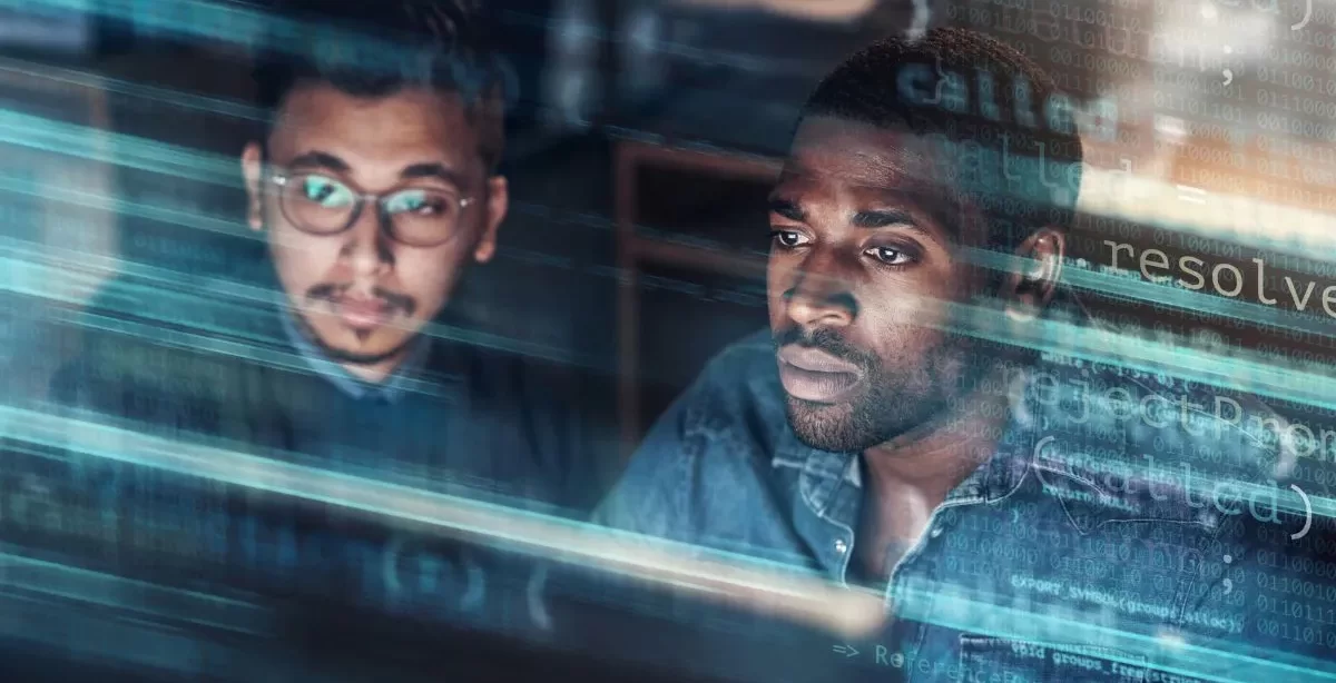 Dois homens, um asiático e outro negro, olham para o mesmo ponto da imagem, provavelmente para a tela de um computador. Sobrepostas a eles temos várias linhas de código.