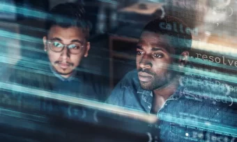 Dois homens, um asiático e outro negro, olham para o mesmo ponto da imagem, provavelmente para a tela de um computador. Sobrepostas a eles temos várias linhas de código.