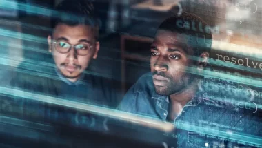 Dois homens, um asiático e outro negro, olham para o mesmo ponto da imagem, provavelmente para a tela de um computador. Sobrepostas a eles temos várias linhas de código.