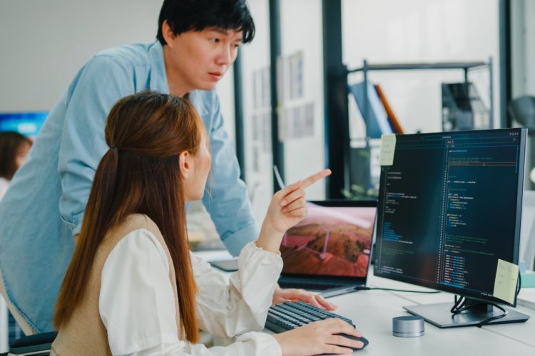 Mulher asiática com blusa amarela e branca parece explicar algo a seu gerente enquanto gesticula. Ele também é asiático, veste uma camisa azul. Olha para a tela de um desktop e parece sério.