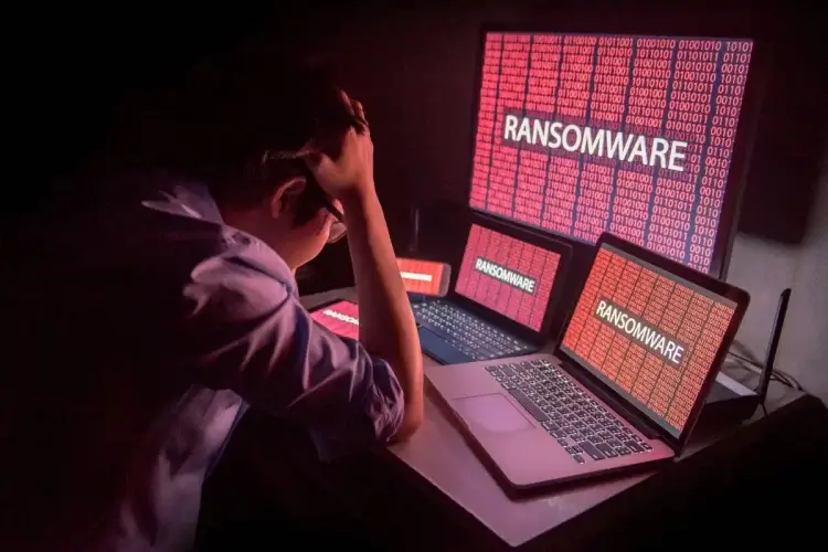Homem de camisa e óculos parece desesperado em frente a várias telas de notebook e um desktop. Nelas, podemos ver códigos binários em vermelho e a palavra "ransomware"