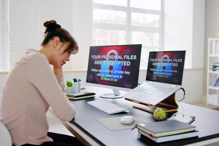 Mulher de coque e blusa rosa está sentada diante de um desktop e um notebook. Ela parece preocupada. Nas telas dos dispositivos, podemos ler em inglês "Your Personal Files Are Encrypted. Make payment or private key will be destroyed in 12 hours 01:34", que em português significa algo como "Seus arquivos estão criptografados. Faça um pagamento ou a chave privada será destruída em 12 horas". 