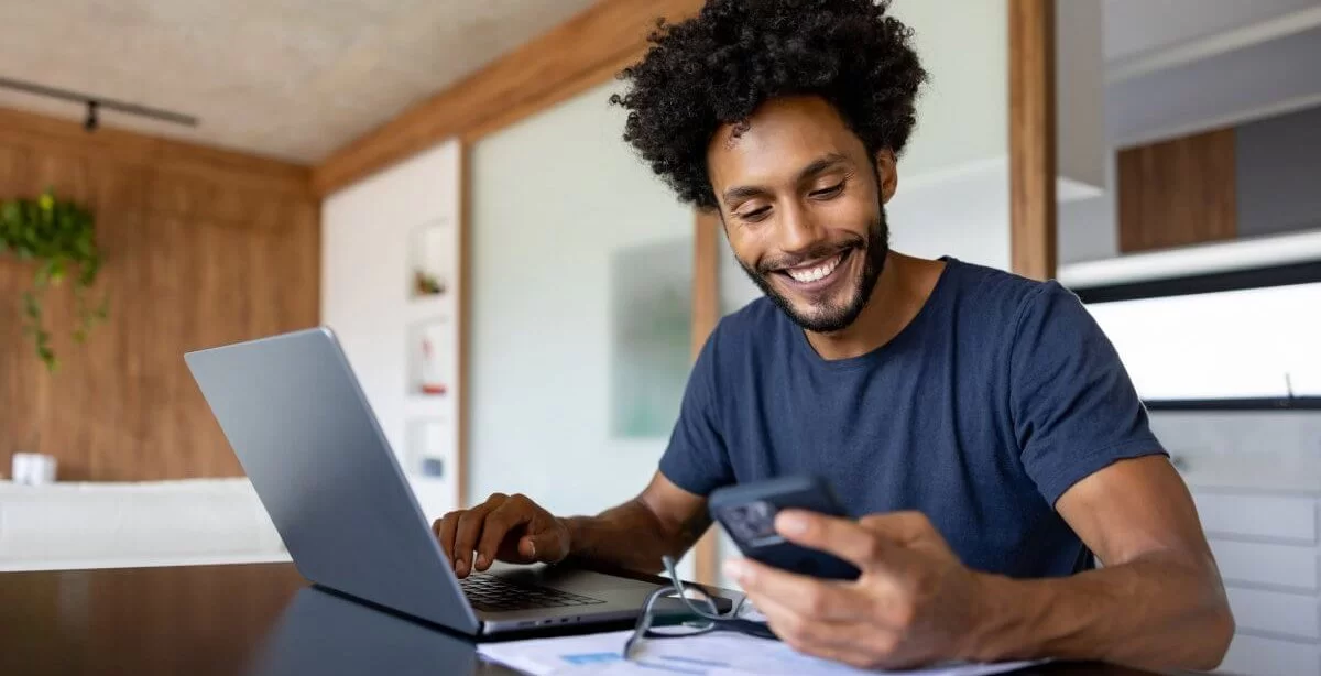 Homem negro sentado diante de um laptop. Ele observa o celular e sorri. Há um par de óculos e papéis em cima da mesa. O ambiente é clean, com detalhes em madeira.