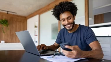 Homem negro sentado diante de um laptop. Ele observa o celular e sorri. Há um par de óculos e papéis em cima da mesa. O ambiente é clean, com detalhes em madeira.