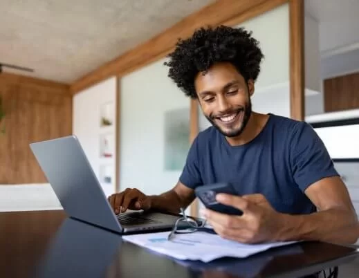 Homem negro sentado diante de um laptop. Ele observa o celular e sorri. Há um par de óculos e papéis em cima da mesa. O ambiente é clean, com detalhes em madeira.