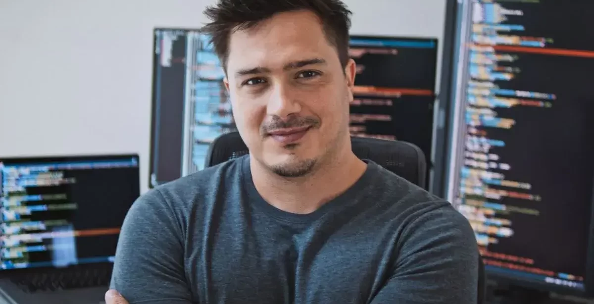 Homem branco, com cabelos escuros, barba e cavanhaque, olha para a câmera com os braços cruzados e sorrindo discretamente. Ele usa uma camiseta cinza. Atrás dele há várias telas de desktop e notebook, que representam o deploy de aplicações no Visual Studio.
