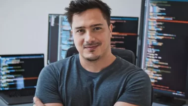 Homem branco, com cabelos escuros, barba e cavanhaque, olha para a câmera com os braços cruzados e sorrindo discretamente. Ele usa uma camiseta cinza. Atrás dele há várias telas de desktop e notebook, que representam o deploy de aplicações no Visual Studio.