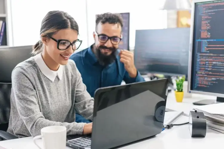 Mulher com cabelo loiro, blusa cinza e óculos olha para a tela de um notebook enquanto sorri. Ao lado dela há um homem com cabelos escuros, barba e óculos, usando uma camisa azul. Ele também sorri enquanto olha para o computador de sua colega de trabalho. O ambiente remete a um escritório, com computadores, canecas, papéis etc. 