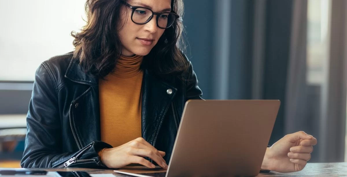 Mulher em ambiente de escritório digitando em um computador com a mão direita. Ela usa óculos, uma blusa cor mostarda e uma jaqueta de couro.