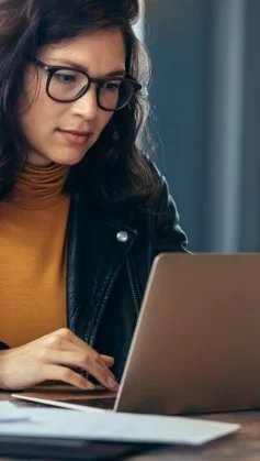 Mulher em ambiente de escritório digitando em um computador com a mão direita. Ela usa óculos, uma blusa cor mostarda e uma jaqueta de couro.