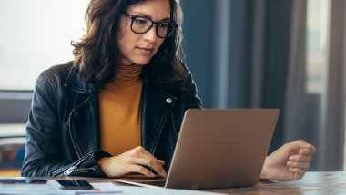 Mulher em ambiente de escritório digitando em um computador com a mão direita. Ela usa óculos, uma blusa cor mostarda e uma jaqueta de couro.