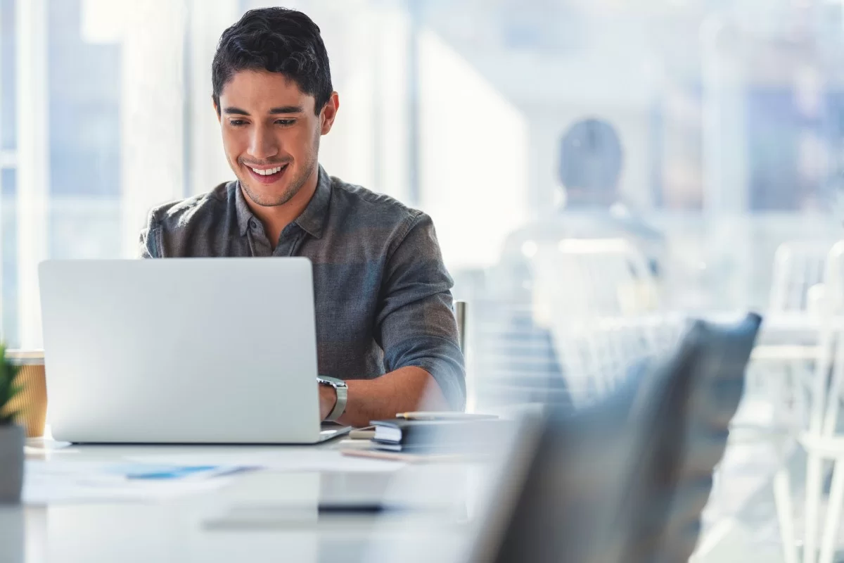 Homem de cabelos escuros e barba sentado em uma cadeira de escritório digitando em um notebook enquanto sorri. 