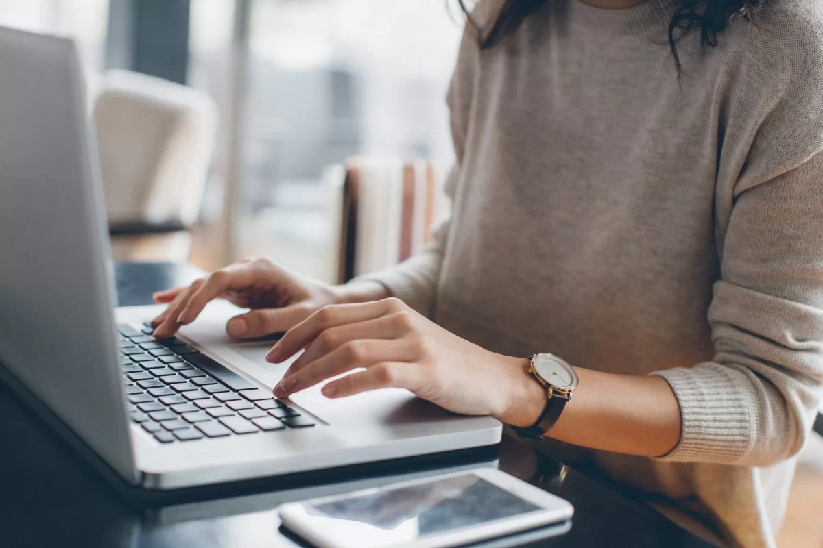 Mulher digitando em um teclado de notebook. Ela usa uma blusa cinza e um relógio no pulso esquerdo. Seu rosto não aparece. Ao lado do laptop há um smartphone. 