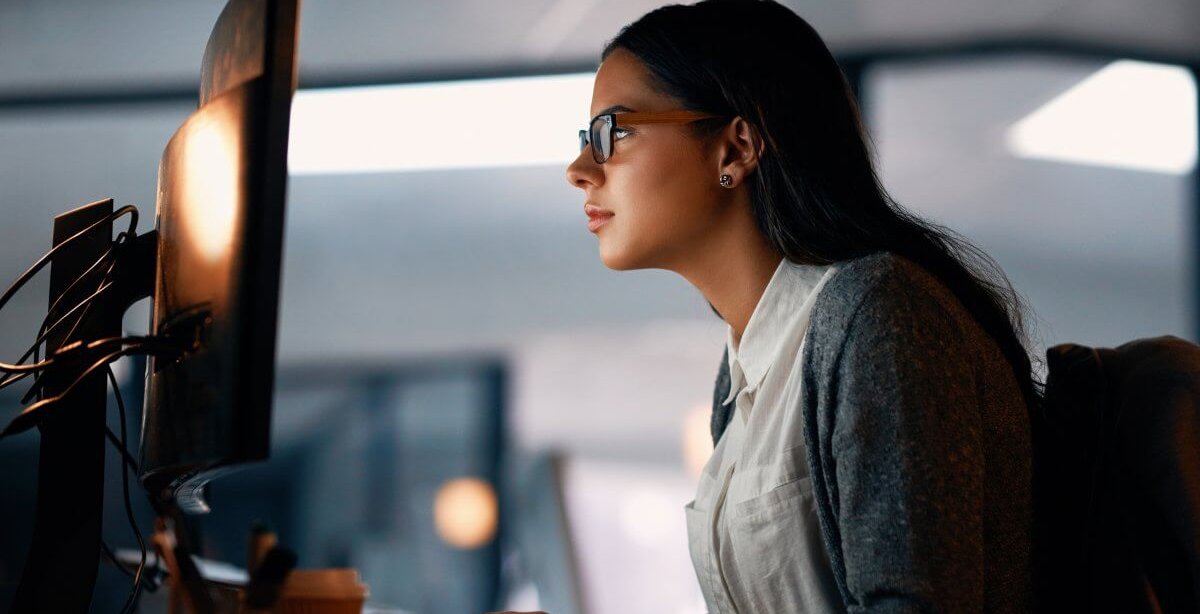 Mulher de óculos digitando em teclado de desktop. A tela não é visível. A mulher está em um escritório, veste camisa branca e casaco azul.