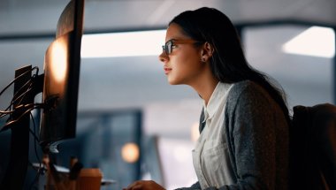 Mulher de óculos digitando em teclado de desktop. A tela não é visível. A mulher está em um escritório, veste camisa branca e casaco azul.