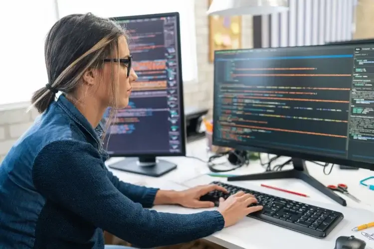 Mulher de óculos com cabelo amarrado, sentada em uma cadeira digitando códigos de Bash em duas telas do computador. Vários códigos aparecem na tela principal e na tela secundária. A cena acontece em um escritório durante o dia.