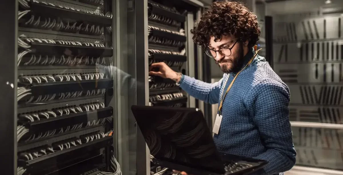 Homem moreno de barba e camisa azul segurando um notebook enquanto faz configurações em um servidor. Ele aparentemente está em uma empresa e usa o notebook para checar algum site para realizar as configurações.