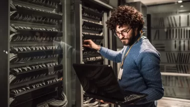 Homem moreno de barba e camisa azul segurando um notebook enquanto faz configurações em um servidor. Ele aparentemente está em uma empresa e usa o notebook para checar algum site para realizar as configurações.