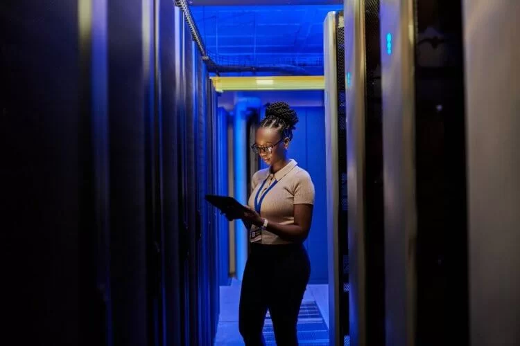 Uma mulher negra está em uma área com servidores. Ela está avaliando como hospedar um site enquanto olha para um tablet. Há diversos corredores com computadores à esquerda e à direita na imagem.