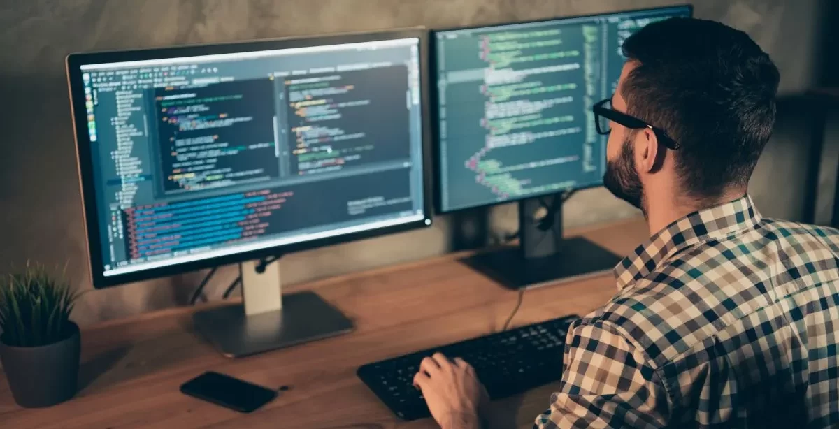 Homem de cabelo escuro, barba, óculos e camisa xadrez trabalhando em dois desktops ao mesmo tempo. Ele está de costas para a câmera. Nas telas, aparecem códigos de programação.