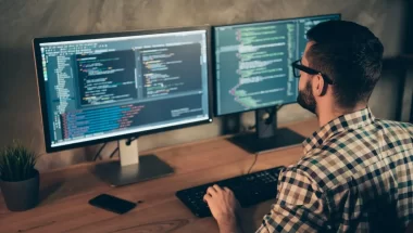 Homem de cabelo escuro, barba, óculos e camisa xadrez trabalhando em dois desktops ao mesmo tempo. Ele está de costas para a câmera. Nas telas, aparecem códigos de programação.