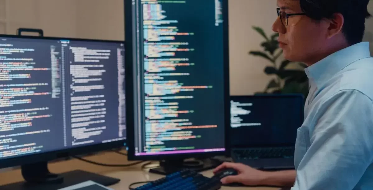 Homem de traços asiáticos, usando óculos e camisa, trabalha com duas telas de computador. Nelas, podemos ver códigos de uma linguagem de programação não identificada.